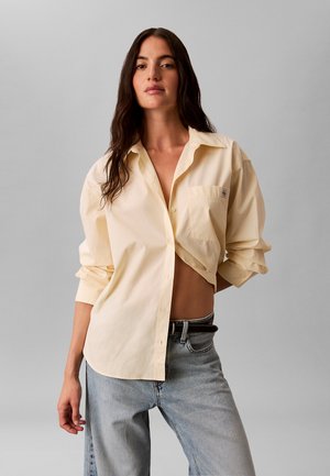 Donna con lunghi capelli scuri che indossa una camicia larga giallo pallido con bottoni, parzialmente aperta, e jeans azzurri con cintura nera.