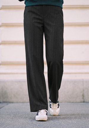 Personne portant un pantalon noir à fines rayures et des baskets blanches marchant sur une surface pavée devant un mur de pierre claire.