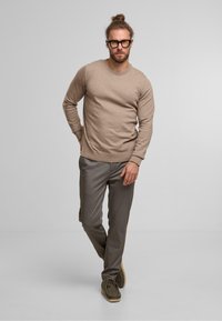 Jersey beige de punto con mangas largas combinado con unos pantalones grises de estampado. El modelo lleva gafas y zapatos marrones. Fondo liso.