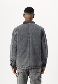 Chaqueta de mezclilla gris con un corte relajado, cuello a presión y mangas largas. Presenta un desvanecimiento sutil y costuras en contraste. Vista trasera.