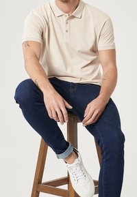 Beige pikétröja med krage och tre knappar, i kombination med mörkblå jeans. Vita sneakers med perforerade detaljer på en träpall.