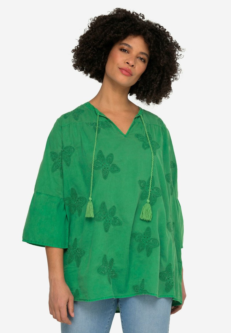 Grøn bomuldsbluse med V-hals, trekvartærmer og blomsterbroderi. Har frynsesnore og et løst, flydende design.