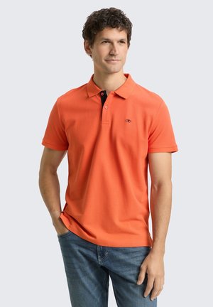 Mężczyzna z kręconymi włosami ubrany w jasnopomarańczową koszulkę polo z krótkim rękawem i niebieskie dżinsy, stoi z jedną ręką w kieszeni na jasnym tle.