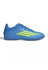 Chaussure de football bleue avec semelle texturée et trois bandes jaunes vives sur le côté, conçue pour les surfaces en gazon synthétique ou turf.
