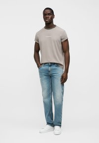 Άνδρας που στέκεται με τα χέρια στις τσέπες ενός ανοιχτό μπλε τζιν, φοράει μπεζ t-shirt με μανίκια γυρισμένα και λευκά αθλητικά παπούτσια σε απλό φόντο.