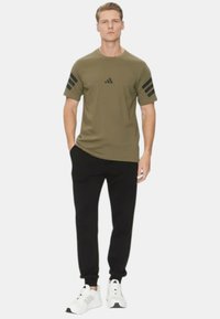 Maglietta sportiva verde oliva con dettagli neri a tre strisce sulle spalle. Pantaloni neri e sneakers bianche completano l'outfit.