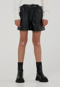 Pantaloni shorts in pelle nera con vita regolabile, caratterizzati da una texture liscia e risvolti. Abbinati a stivali neri alla caviglia con dettagli sottili sulle ginocchia visibili.