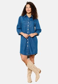 Denim-paitapusero, jossa on napitettava etuosa, rintataskut ja puolipitkät hihat. Yhdistetty vaaleanbeigeihin polvipitkiin saappaisiin.
