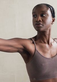 Sutiã desportivo castanho com uma larga faixa inferior e alças, apresentando uma textura suave. O modelo está numa pose com os braços estendidos.