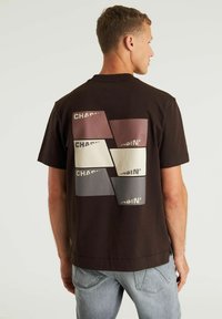 Korte mouwen, donkerbruine t-shirt met een grafisch ontwerp op de achterkant met overlappende rechthoekige patches in gedempte kleuren en tekstaccenten.