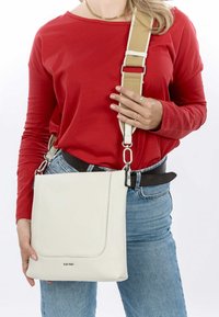 Bílá kožená crossbody taška s hladkým povrchem, s klopovým uzávěrem, nastavitelným béžovým popruhem a kovovými akcenty.