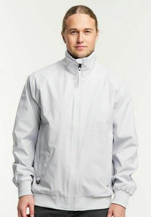 Volwassen man met lang haar die een witte jas met rits en een hoge kraag draagt, staand tegen een effen lichte achtergrond.