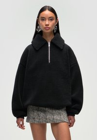 Czarny polarowy sweter z kołnierzem i półzapinem z przodu. Posiada oversized rękawy i puszystą teksturę. Noszony na wzorzystą mini spódnicę.