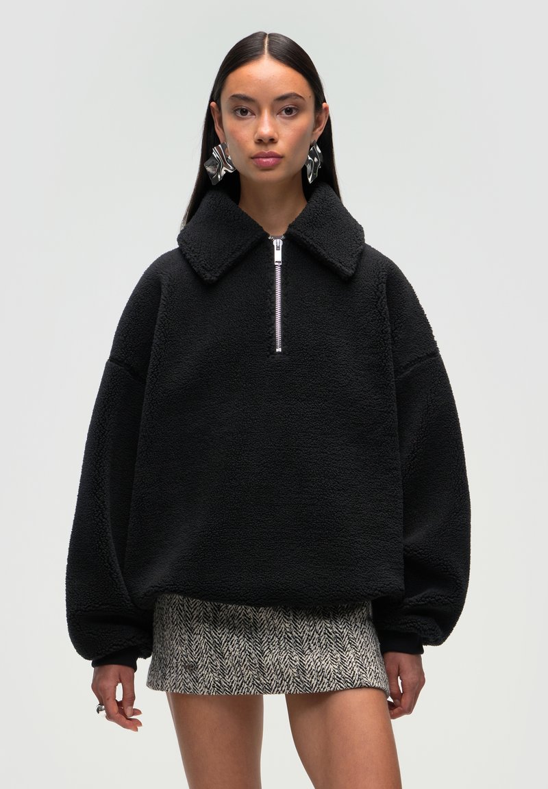Czarny polarowy sweter z kołnierzem i półzapinem z przodu. Posiada oversized rękawy i puszystą teksturę. Noszony na wzorzystą mini spódnicę.