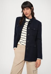 Tengerészkék blézer álló gallérral, két elülső zsebbel és gombos záródással. Egy krémszínű, fekete csíkos pulóver fölött, bézs nadrággal viselve.