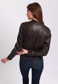 Donna con lunghi capelli castani, che indossa una giacca di pelle marrone scuro e jeans blu, girata leggermente di lato, che sistema la manica della giacca.
