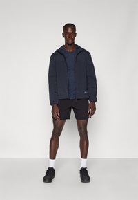 Sort puffer jakke med hætte, båret over en marineblå t-shirt og sorte shorts. Har elastiske manchetter og lynlås. Sorte sneakers og hvide sokker.