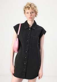 Zwarte denim shirtjurk met korte mouwen, knoopsluiting aan de voorkant, gebogen zoom en een borstzak. Vergezeld door een kleine roze tas.