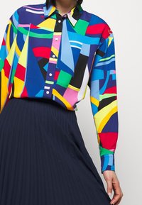Kolorowa, koszula w abstrakcyjny wzór z długim rękawem, z zapięciem na guziki z przodu oraz kontrastowym kołnierzykiem. Połączona z plisowaną, granatową spódnicą.