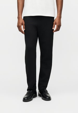 Mężczyzna ubrany w czarne spodnie o prostym kroju, czarne skórzane buty oraz białą koszulę, stojący na tle jasnego, jednolitego tła.