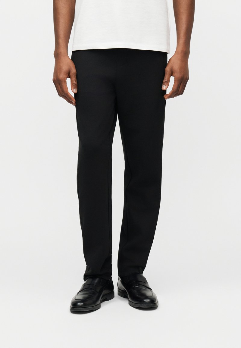 Άνδρας που φοράει μαύρο παντελόνι κανονικής γραμμής, μαύρα δερμάτινα παπούτσια και λευκό πουκάμισο, στέκεται μπροστά σε απλό φωτεινό φόντο.