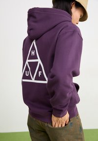 Haut à capuche violet en tissu doux arborant un grand logo triangulaire blanc avec les lettres "H," "U," et "F," porté sur un pantalon marron clair.