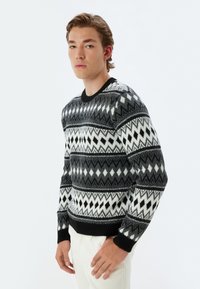 Jersey de patrón en blanco y negro con diseños geométricos, cuello redondo, puños y dobladillo acanalados. Lleva encima un pantalón de color claro.