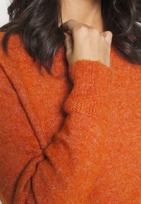 Jasnopomarańczowy sweter wykonany z miękkiego, teksturowanego materiału z okrągłym dekoltem. Posiada ściągacze na mankietach i luźny krój.