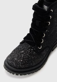 Botín negro con parte superior brillante, cordones de terciopelo y ojales metálicos redondos. Presenta un diseño texturizado y una suela de goma resistente.
