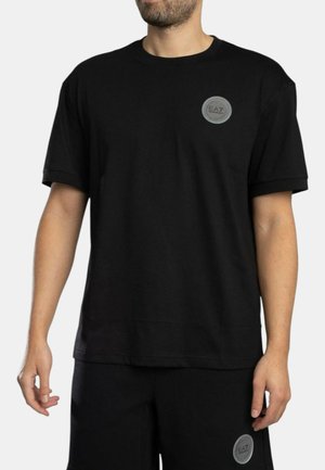 Uomo che indossa una T-shirt nera a maniche corte e pantaloncini, entrambi con il logo circolare grigio EA7 sul petto sinistro e sulla coscia sinistra rispettivamente.