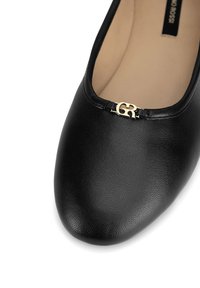 Bailarina de cuero negro con punta redondeada, superficie texturizada y detalle de logo con acento dorado en la abertura frontal.