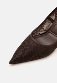 Chaussure plate à bout pointu avec dessus en dentelle florale brun foncé et bord assorti, montrant une semelle intérieure matelassée.