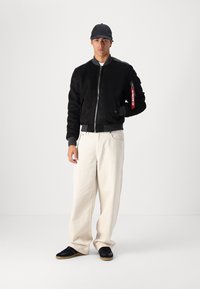 Giubbotto bomber nero con zip frontale e un patch rosso sulla manica, abbinato a pantaloni beige larghi e sneakers nere.