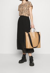 Top s hnědým vzorem, černá plisovaná sukně, černé kotníkové boty a béžová taška s černými rukojeťmi. Zaměřen�í na elegantní současný design a barevný kontrast.