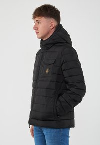 Giacca a vento nera con cappuccio, caratterizzata da una texture trapuntata, chiusura frontale con zip e tasca sul petto con patch logo.
