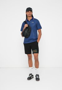 Marineblåternet polo-shirt med gul krave, sorte shorts med et "Riviera" grafik, sort crossbody taske og sorte sneakers.
