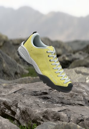 Rumena pohodniška čevlja s sivimi vezalkami in podplatom, v zraku nad skalnatim terenom z zamegljenimi gorami v ozadju.