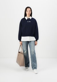 Marineblauwe sweatshirt met wit logo, gelaagd over een wit overhemd met kraag, gecombineerd met lichtblauwe versleten jeans en een beige handtas. Witte sneakers.