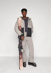 Άνδρας φορώντας σκιώτο, παντελόνι, μπότες και γυαλιά σκι, κρατώντας σκι όρθια σε λευκό φόντο.