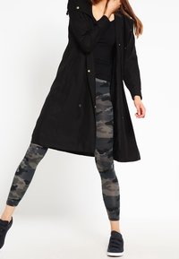 Donna in un lungo cappotto nero, top nero, leggings grigi mimetici e sneakers nere, che sta in piedi con una gamba estesa di lato.