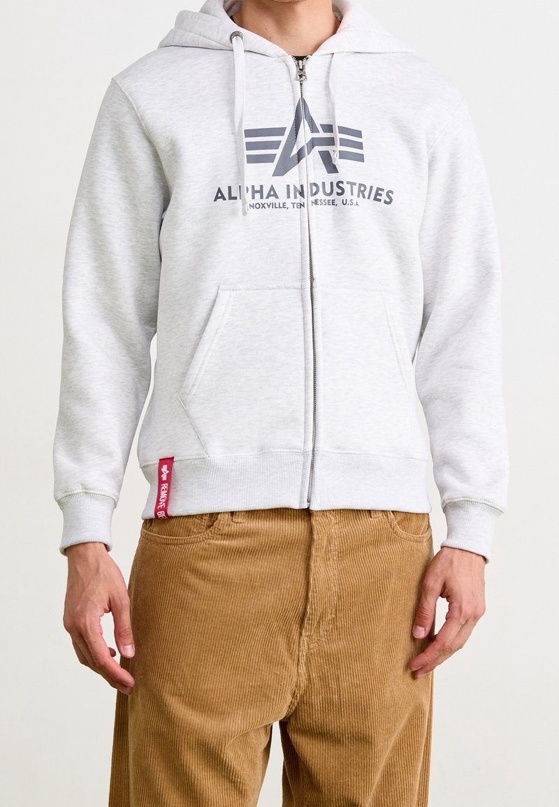 Homme portant un sweat à capuche zippé Alpha Industries gris clair et un pantalon en velours marron, les mains détendues le long du corps.