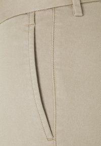 Béžové kalhoty s hladkým povrchem, které mají boční kapsu a jemné diagonální pruhy ve tkanině. Šití na detailech v pase.
