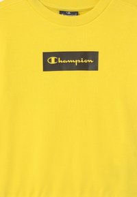 Felpa gialla realizzata in tessuto testurizzato, con un patch rettangolare nero al centro, recante la scritta "Champion" in carattere corsivo giallo.