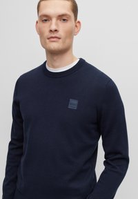 Navyblauwe trui met een ronde hals, ribgebreide manchetten en een klein vierkant logo-patch op de borst. Glad textuur, slim fit ontwerp.