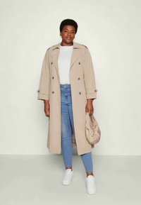 Beige trenchcoat med doble knapper, oppbrettede ermer, iført lyseblå jeans og hvite sneakers; bærer en kremfarget veske.