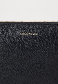 Poche en cuir noir avec surface texturée, fermeture éclair dorée et logo "Coccinelle" en relief. Forme compacte et rectangulaire.