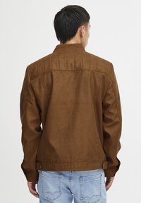 Veste marron en suédine avec un col structuré, des coutures verticales au dos et des boutons pression à l'ourlet. Portée avec un jean bleu clair.