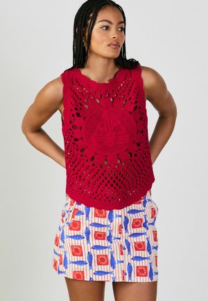 Frau mit geflochtenem Haar, die ein ärmelloses rotes Häkeloberteil mit Segelboot-Motiv und einen gemusterten Rock mit blauen Fischen und roten Quadraten trägt.