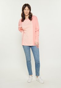 Maglietta a maniche lunghe rosa chiaro con un design a pannello testurizzato, abbinata a jeans skinny blu e sneakers bianche su uno sfondo neutro.