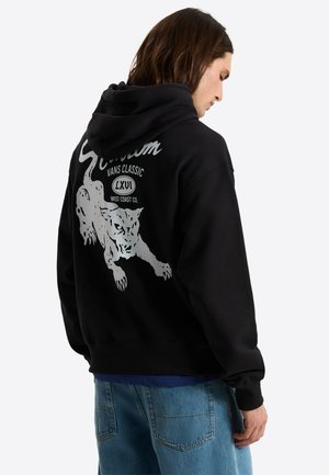Młody mężczyzna w czarnej bluzie z kapturem z białą grafiką skaczącego lamparta i napisem "VANS CLASSIC LXVI WEST COAST CO." na plecach.